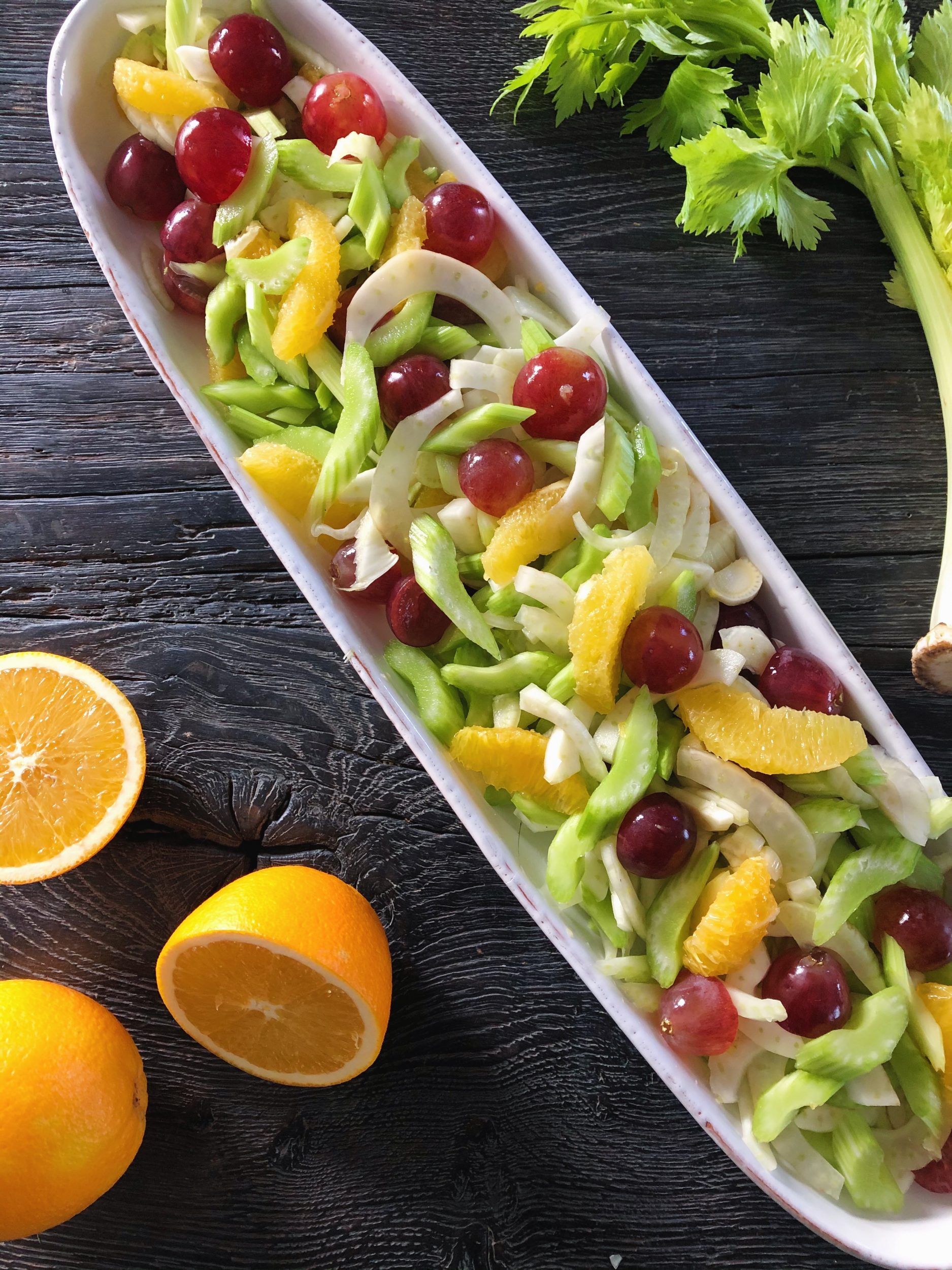 Et aflangt hvidt keramikfad ses på et sort rustikt træbord. I fadet er der en farvering appelsinsalat med vinduer i forskallige farver. Der ses også et bundt bladselleri ligge ved siden af fadet. På bordet ligger også tre halve appelsiner.