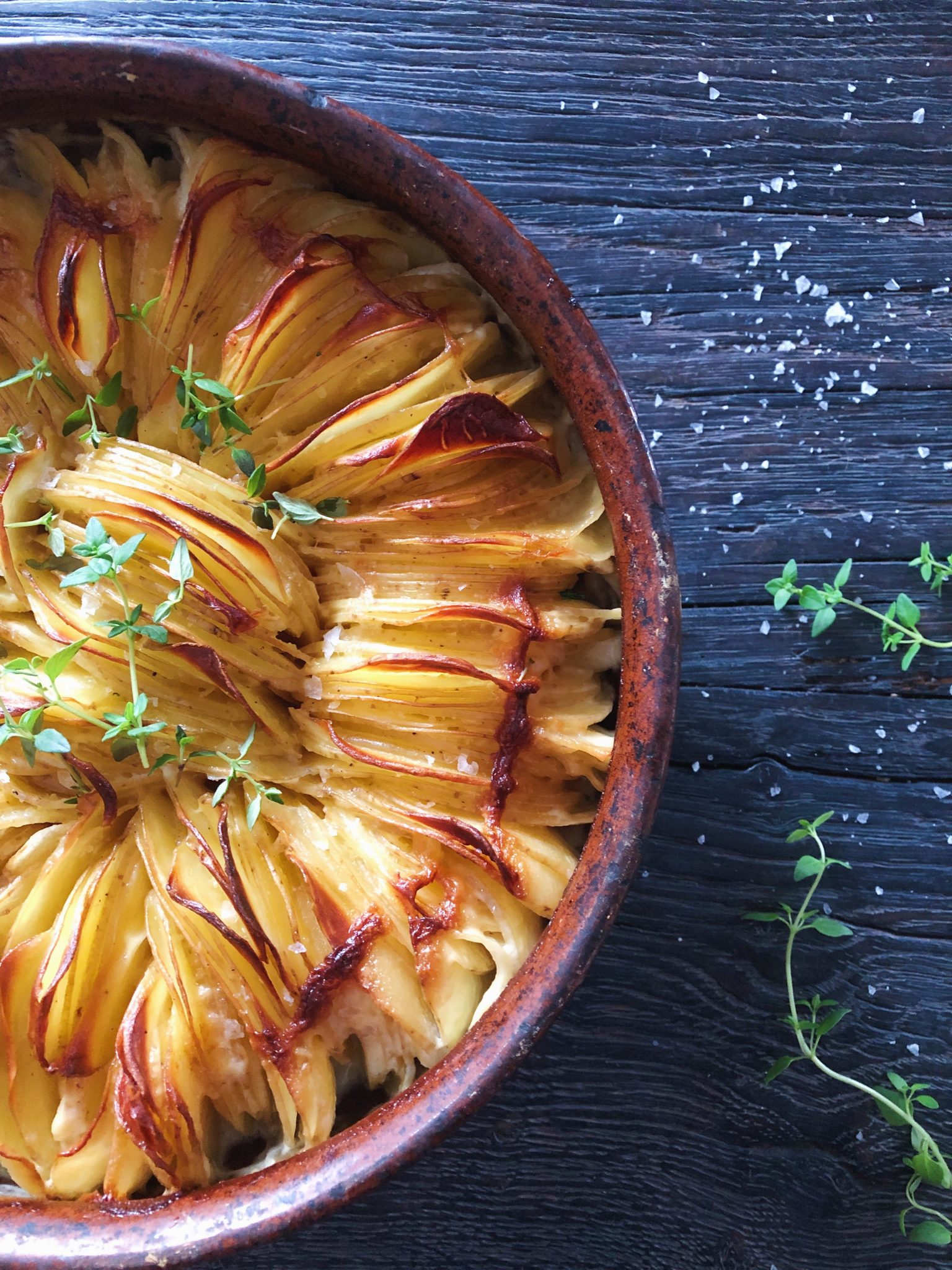 Flødekartofer der er delvist sprøde (den del der er synlig af kartoffelskiverne). De ses i et fad med kartofler i skiver på højkant der ser bagte ud. Fadet står på et sort rustikt træbord med et par kviste timian liggende ved siden af