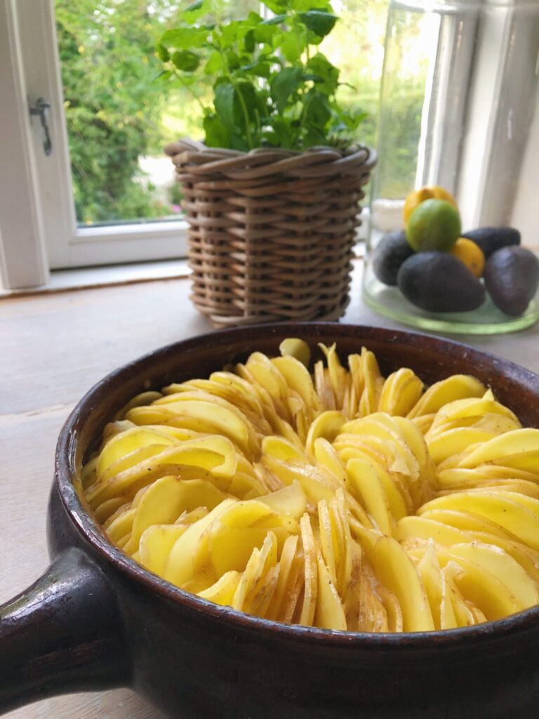 Kartofler skåret i skiver og placeret på højkant i brunt fad inden de skal i ovnen. Det er flødekartofler med sprød kant. I baggrunden ses flettet rund kurv med grøn krydderurt i