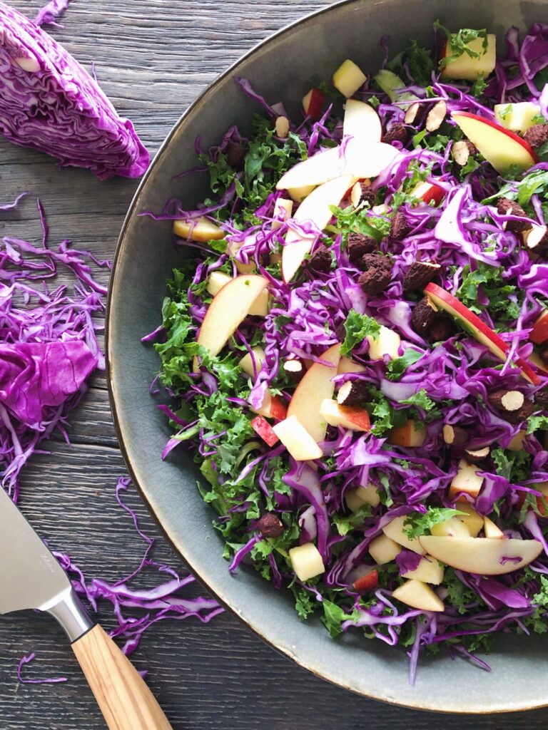 kålsalat med sprøde soyamandler ses i et gråt fad. Der er brugt rød spidskål i salaten. Der ligger noget udskåret spidskål ved siden af fadet. Man skimter ligeledes en Saladine grøntsagskniv