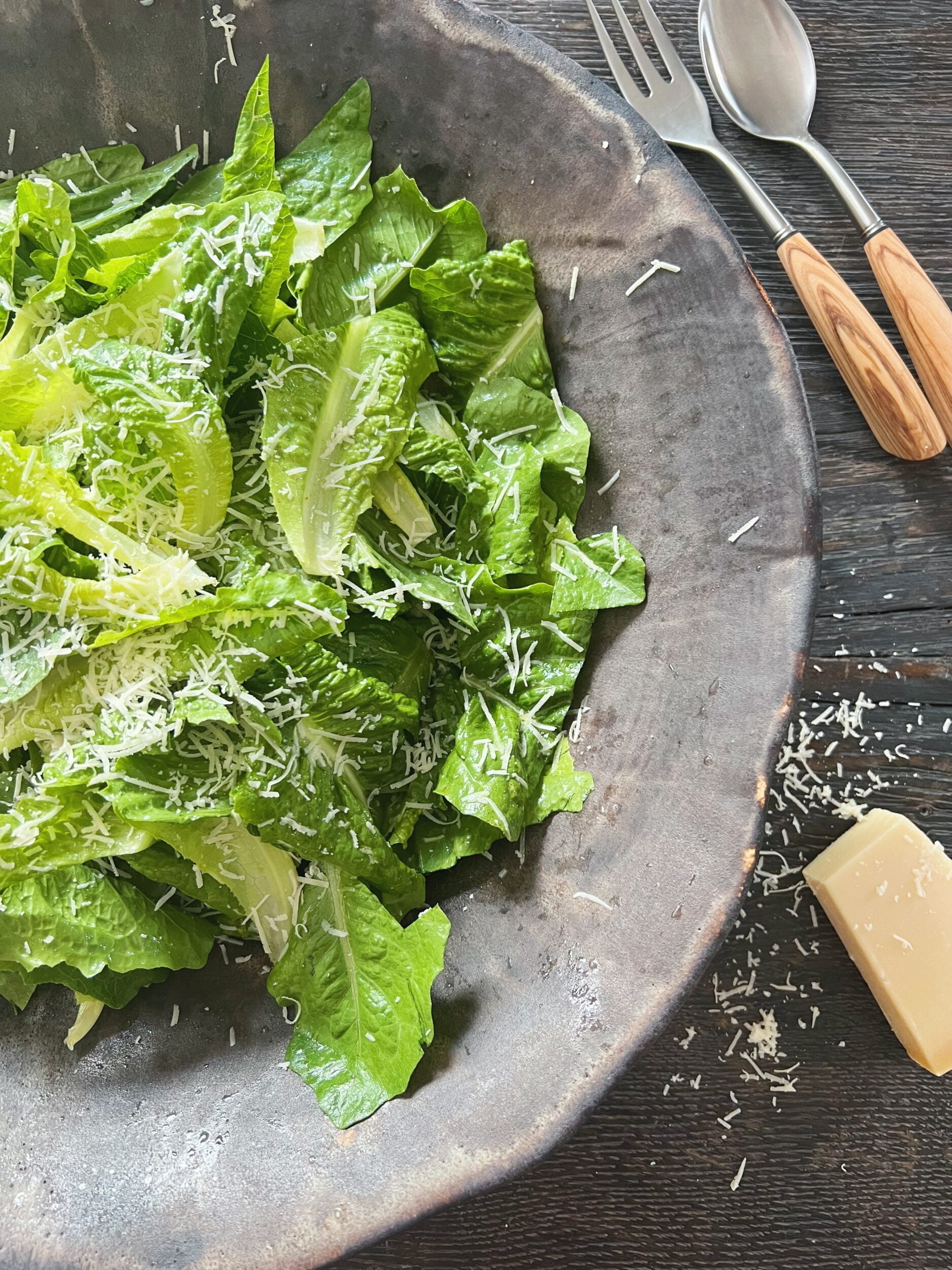 sort rustik keramikskål med romainesalat der er toppet med revet parmesanost ved siden af ses salatbestik og parmesan på bordet