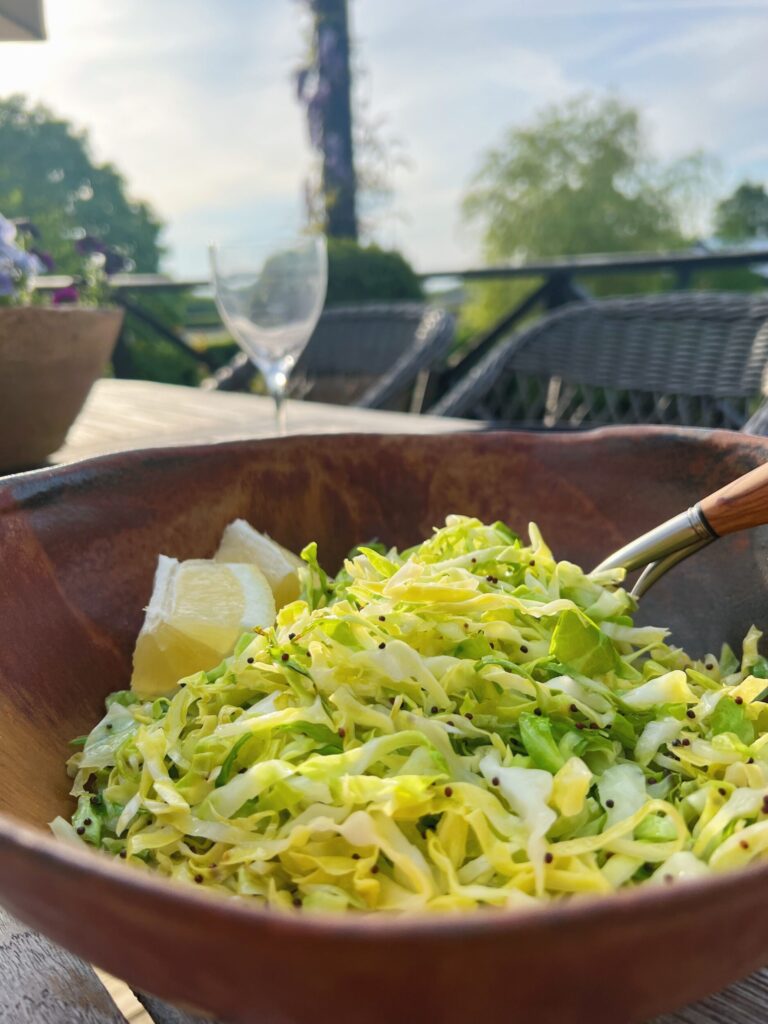 lun spidskålssalat med brune sennepsfrø og citronsaft i rustbrun salatskål sammen med to salatbåde sat på et bord udenfor i baggrunden fornemmes himmel og natur