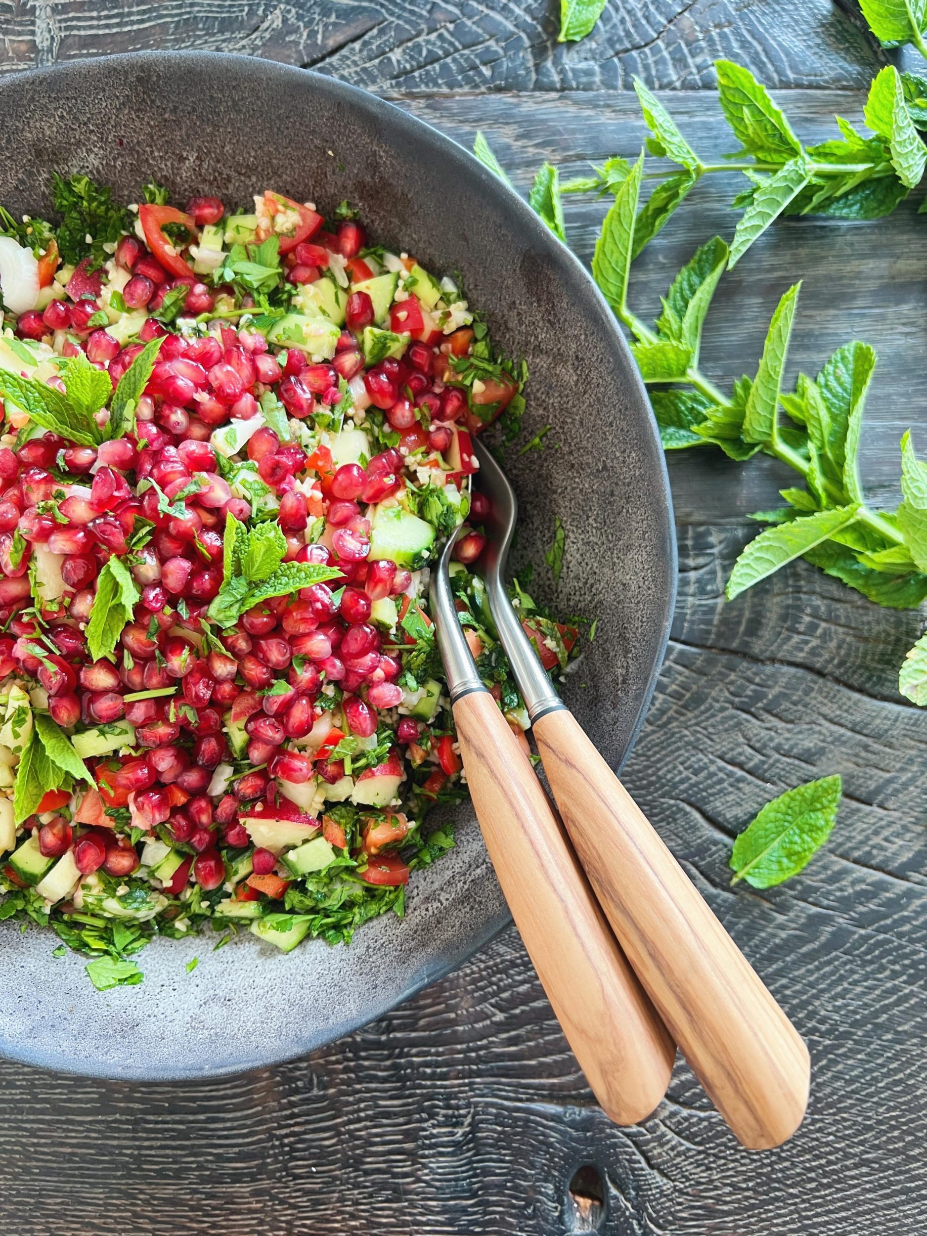 På et sort bord ses en farverig salat i en sort blackish salatskål. Det er en bulgursalat med ferskner etc. Der er et salatbestik stukket i salaten. Det har skaft af oliventræ. Salatskålen står på et sort træbord og der ligger stilke af mynte ved siden af.