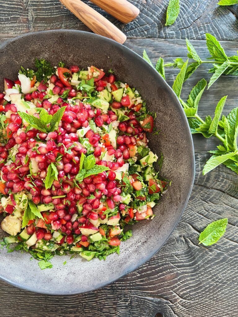 På et sort bord ses en farverig salat med bulgur i en sort blackish salatskål. Salatskålen står på et sort træbord og der ligger stilke af mynte ved siden af og salatbestik skimtes