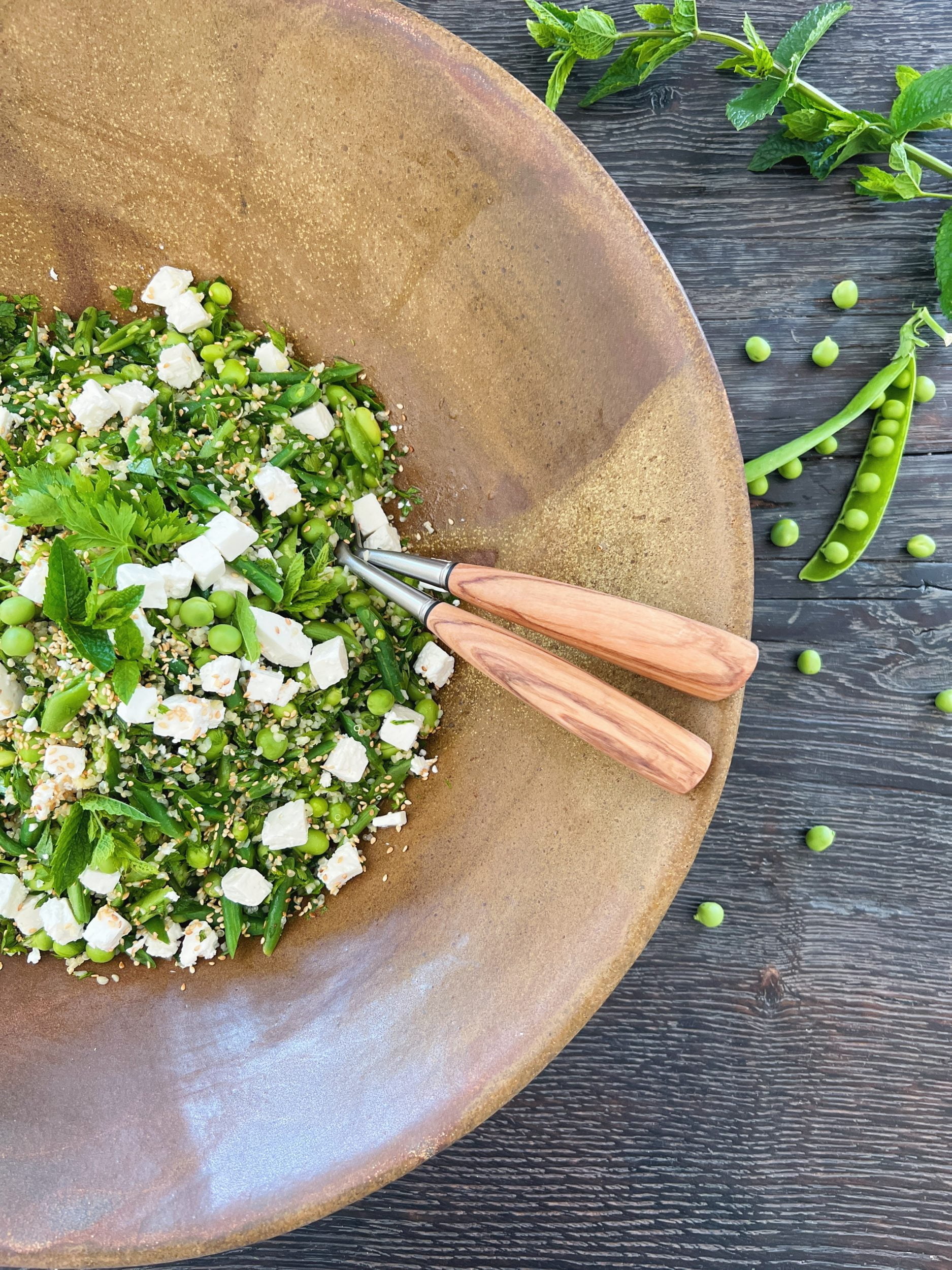 stort brunt fad med en quinoasalat i sammen med ærter bønner slikærter feta og sesamfrø. Der er bestik i fadet og det står på et sort træbord