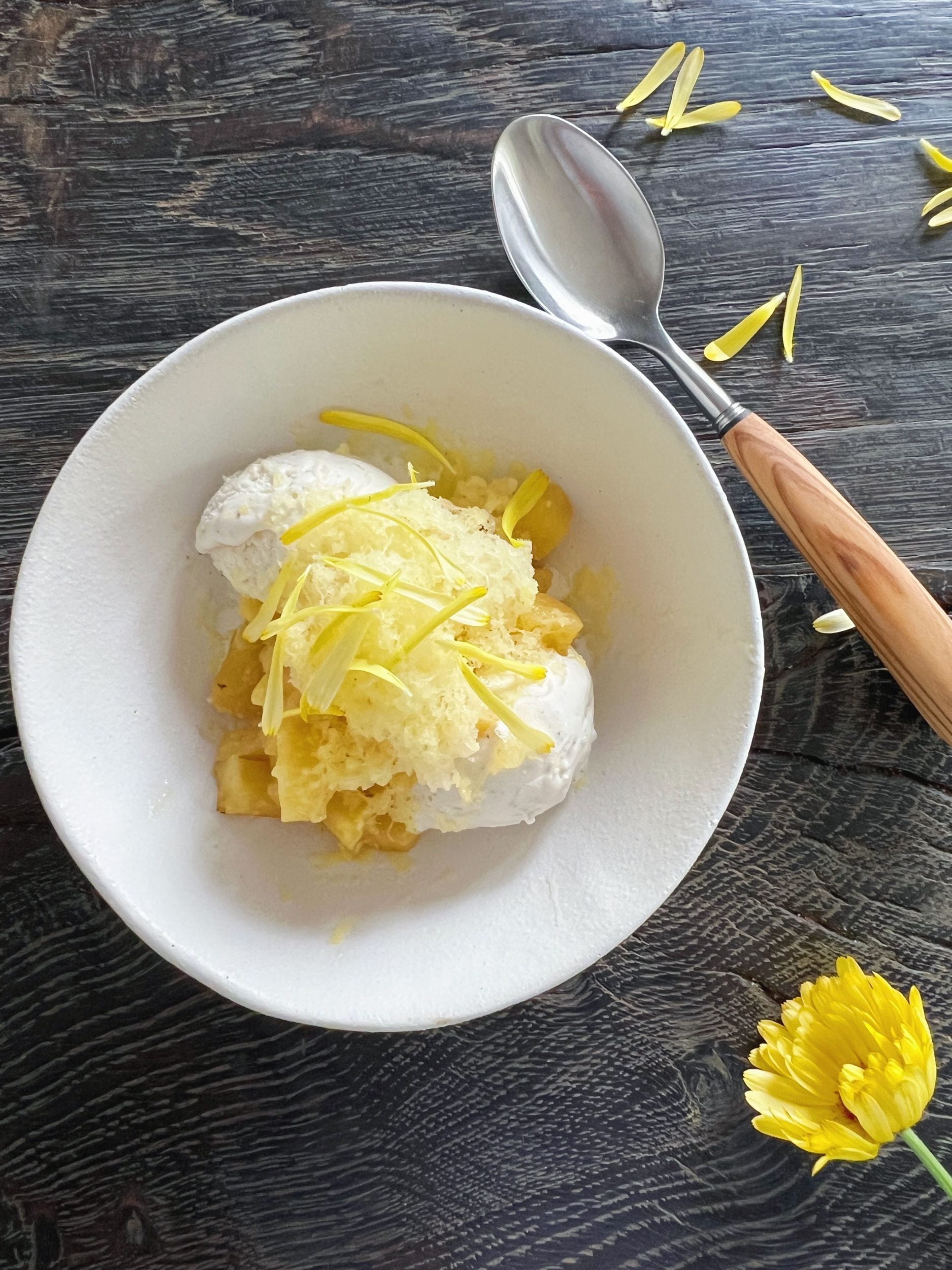 dessert med ananas og is pyntet med gule kronblad fra blomst i hvid skål sat på sort rustikt træbord hvor der også ligger en gul blomst