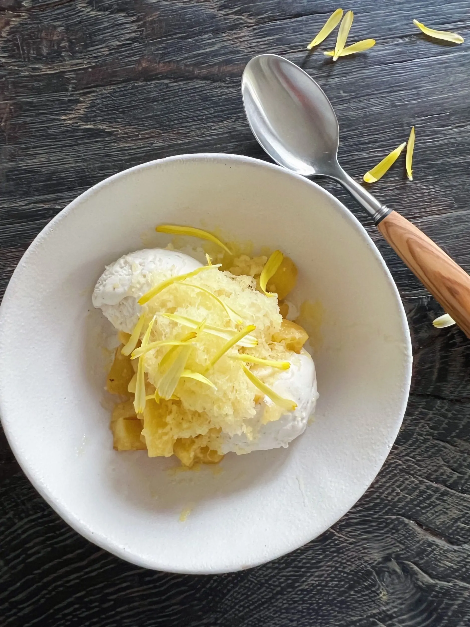 dessert med ananas og is pyntet med gule kronblad fra blomst i hvid skål sat på sort rustikt træbord hvor der også ligger en dessertske med skaft af oliventræ