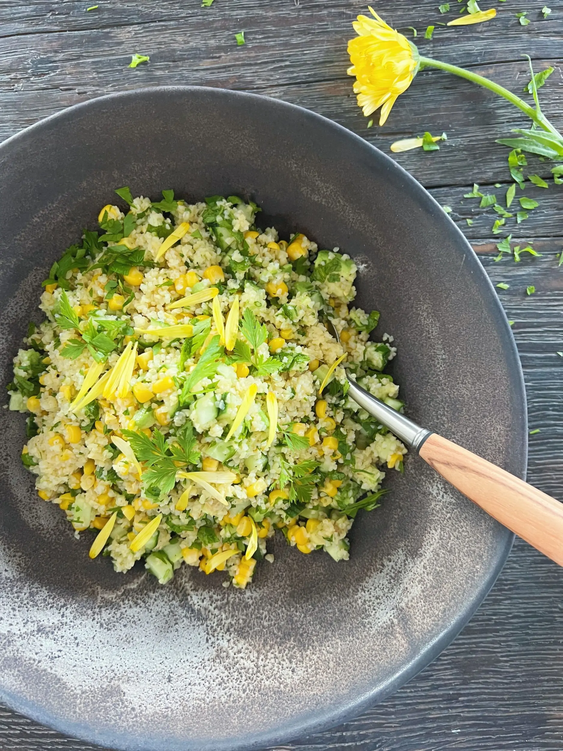 bulgursalat med majs i gråbrun keramik salatskål med en ske i med oliventræskaft. Pynter med gule blomster.
