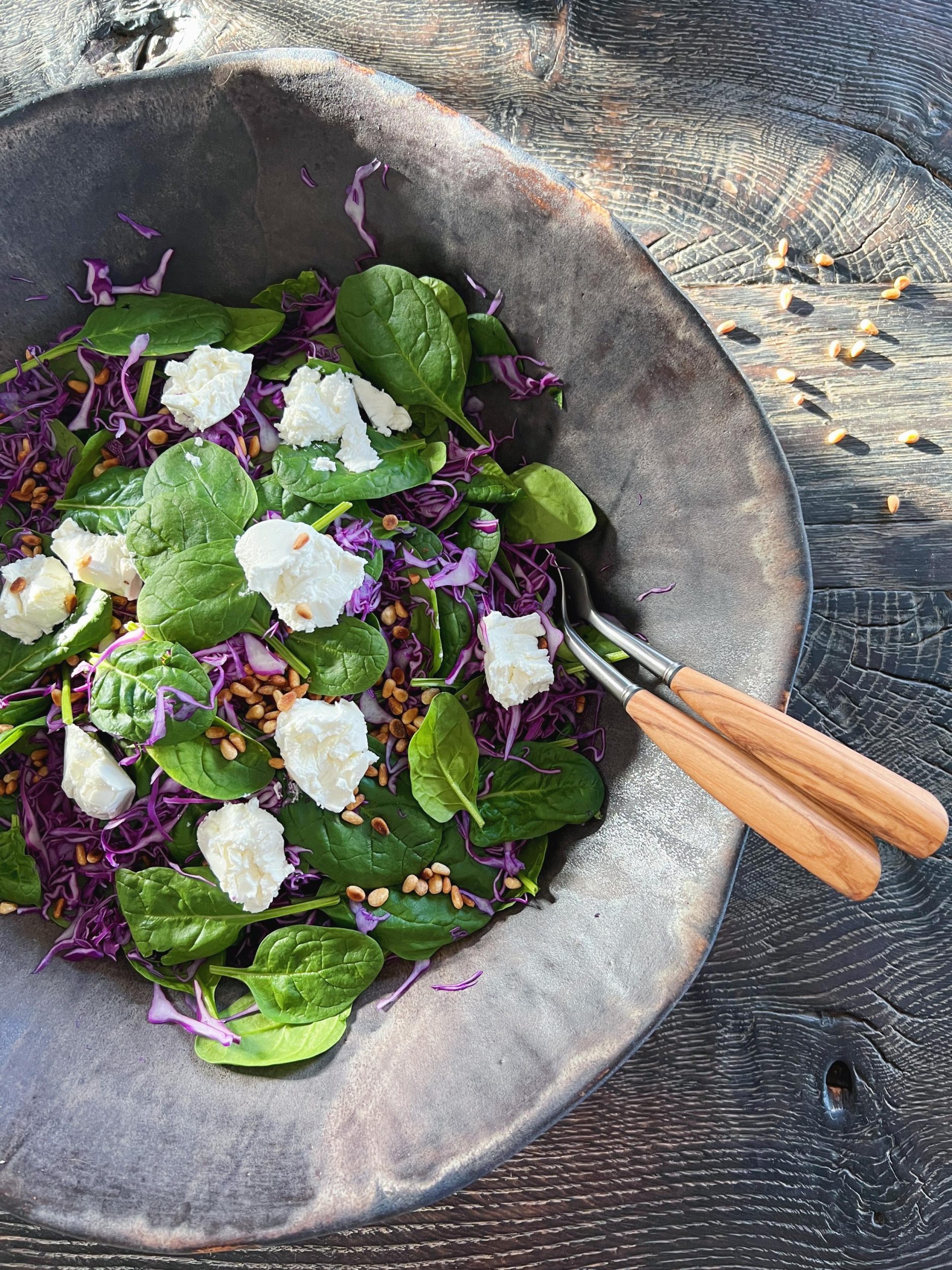 spidskålssalat med spinat, ristede pinjerkerner og cremet gedeost ses i stort rustik keramikfad. Der er placerer et saladinie salatbestik i fadet. Der ses stråler af solskin der passerer ved siden salatskålen, der står på et sort rustikt træbord.