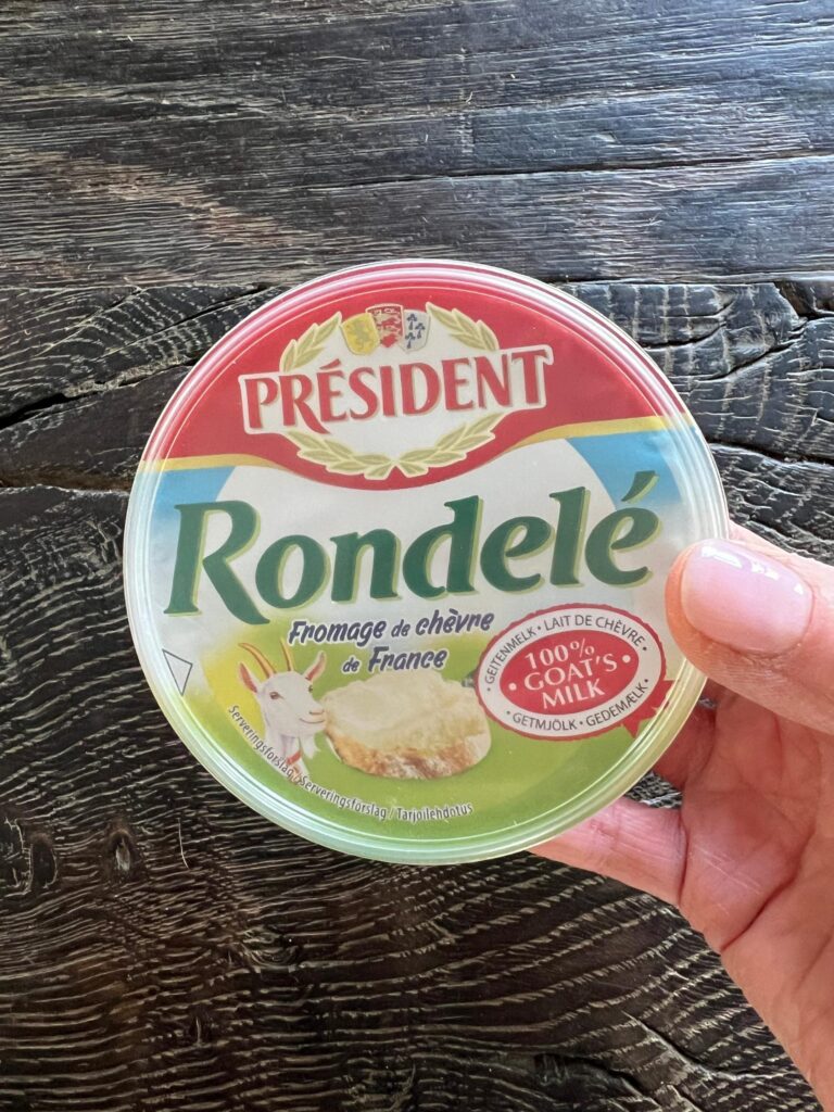 Ost brugt til lun olivensnack. Gedeosten président rondele chévre i plastemballage ses holdt i hånden. I baggrunden ses et sort rustik træbord. Emballagen er i røde, blå og grønne nuancer og der er en ged afbilledet