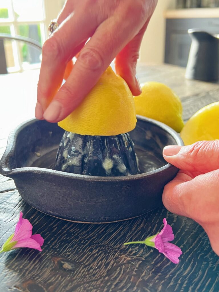 En hånd trykker en overskåret citron ned over en citronpresse i sort keramik. I forgrunden ses to lyserøde blomsterhoveder der ligger på bordet. Bordet er et sort træbord. I baggrunden fornemmes et køkken med sorte fronter.