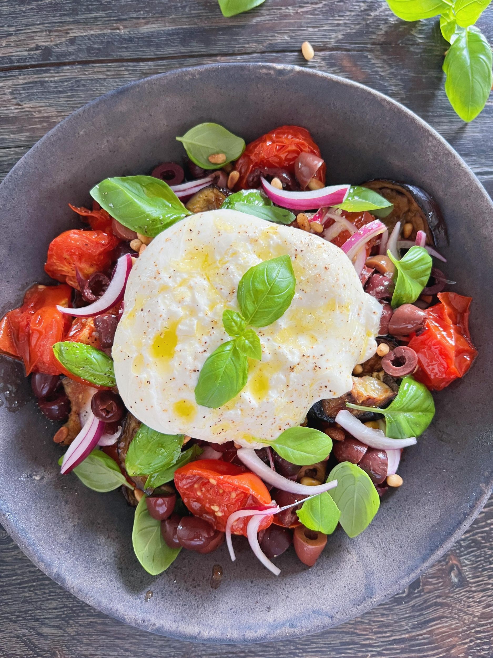 en salat ses tæt på. Der er tomater, auberginer, rødløg og basillikum i. På toppen ligger en hel burrata ost der er pyntet med basilikumblad.