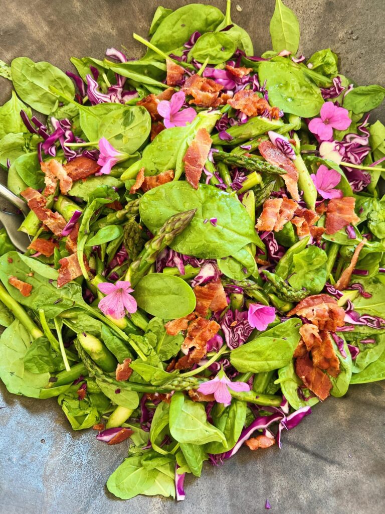 En salat ses helt tæt på. Den er serveret i et sort keramik fad. Det er en spinatsalat, da der ses en masse spinat og rød kål. Der ligger sprød bacon på toppen og salaten er pyntet med lyserøde blomster, kaldet storkenæb.
