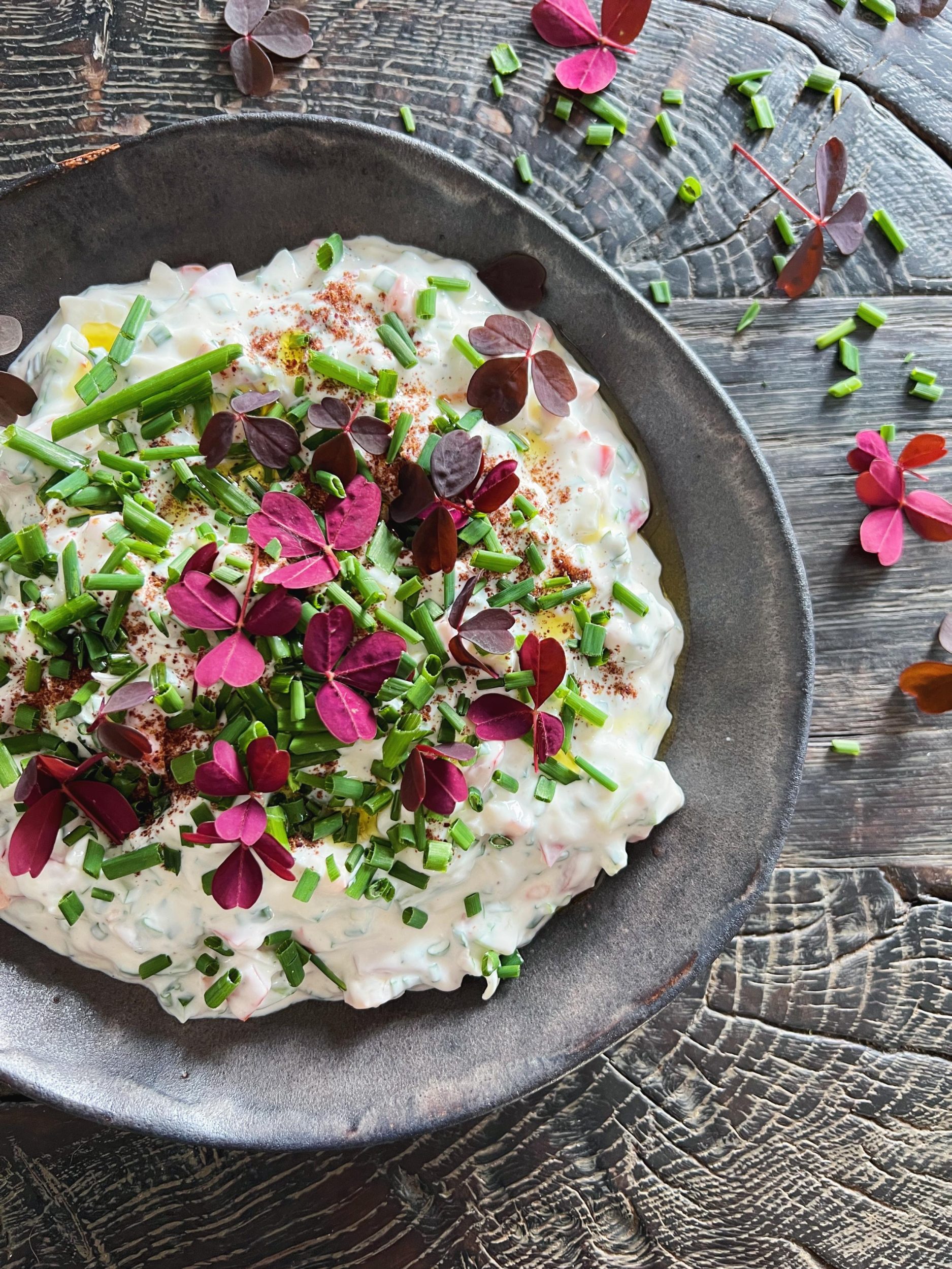 en salat med yoghurtdressing ses serveret i en sort rustik keramikskål. Skålen står på et sort rustikt træbord. Salatopskriften er toppet og pyntet med klippet purlig og blade fra rød skovsyre. På bordet ligger lidt afklip fra purløg og røde blade fra skovsyre