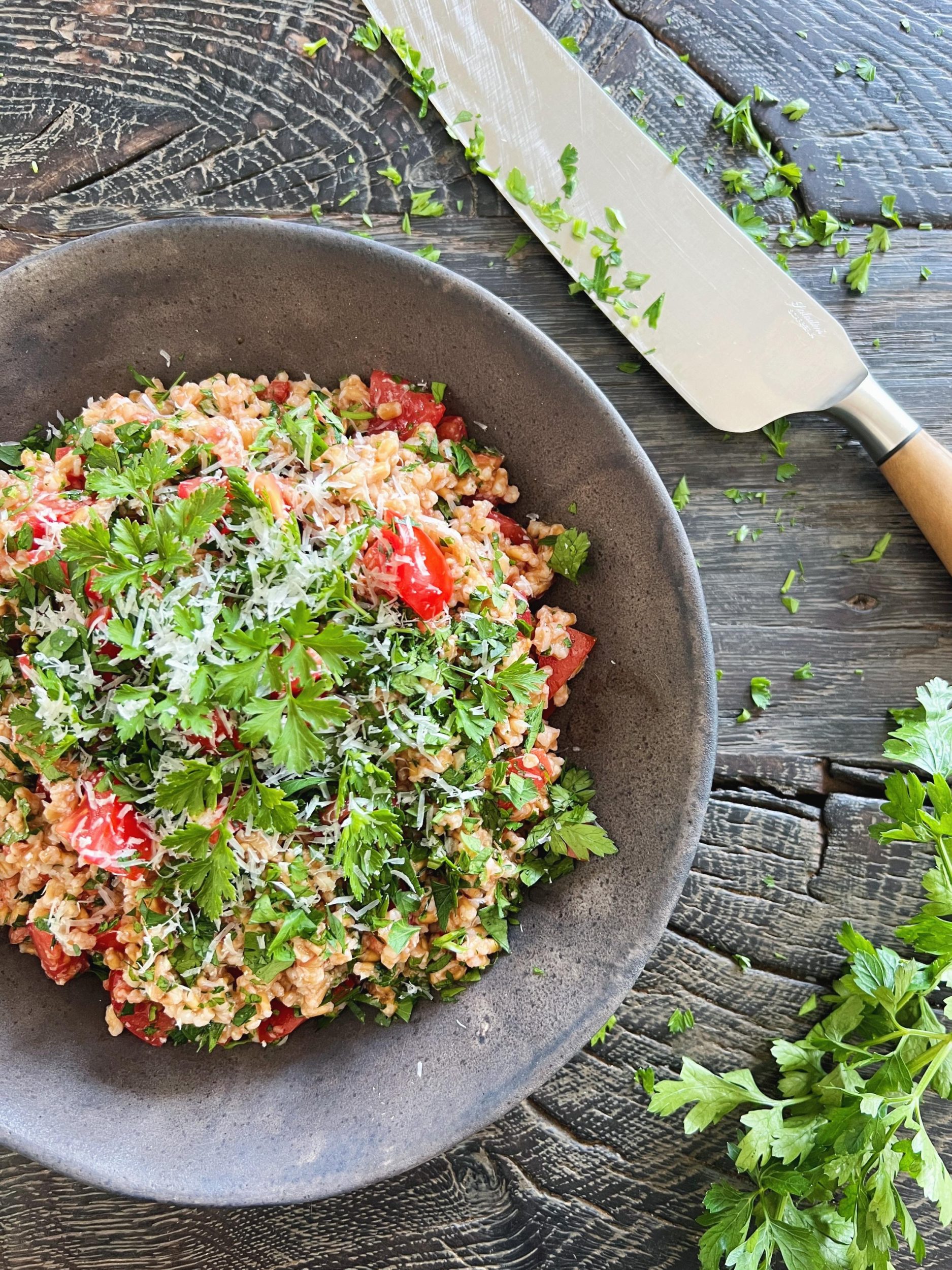 En speltkernesalat med persille, parmesan og tomatdressing ses serveret i en rund rustik sort keramikskål. Den er toppet med hakket bredbladet persille. Den står på et sort rustikt sort træbord. Ved siden af ligger en santukokniv med rester af persille på. Den har skaft i oliventræ.