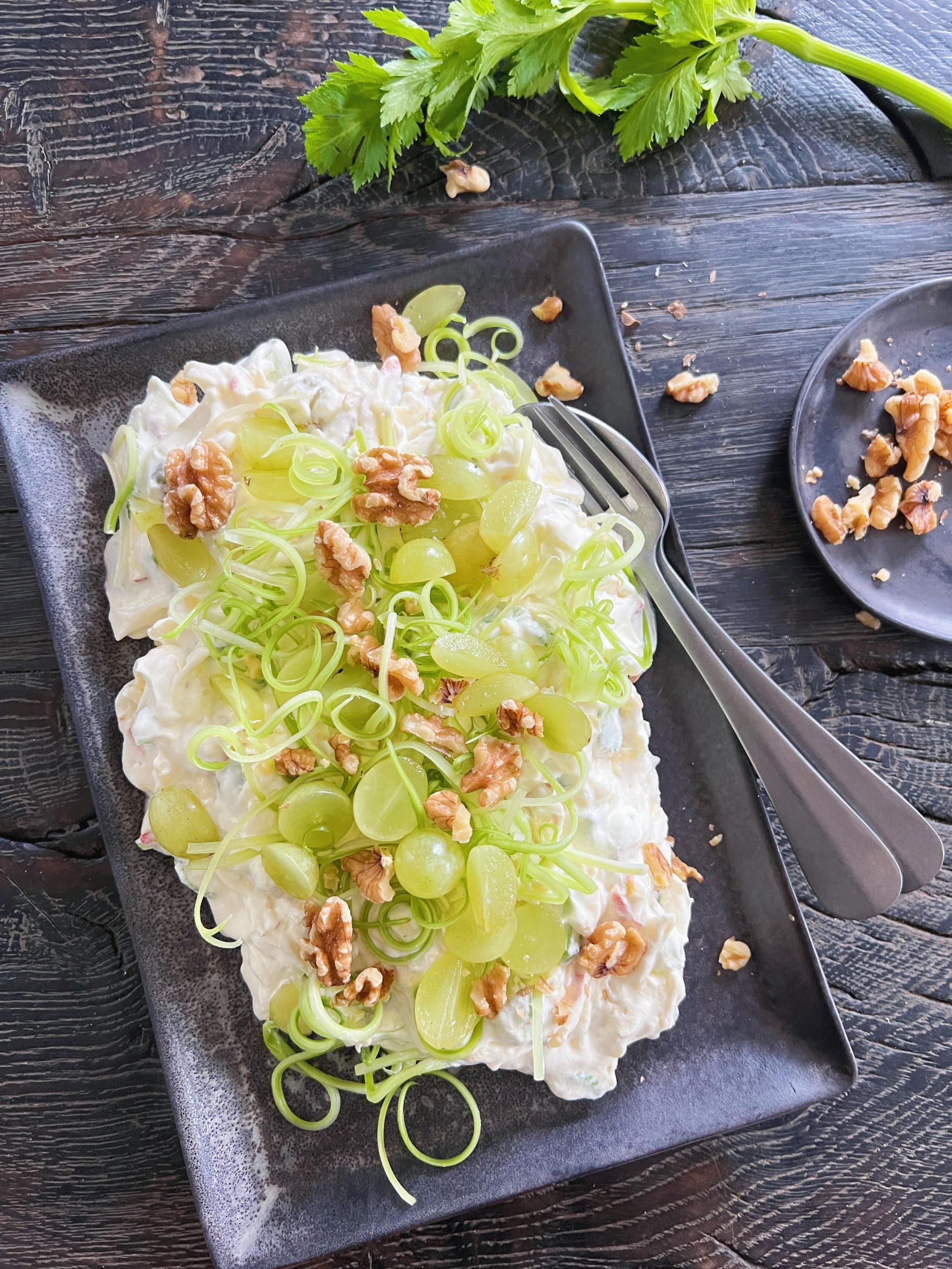 En waldorfsalat ses anrettet på et firkantet sort keramikfad fra brandet BadAss Ceramics. Den klassiske waldorfsalat er toppet med vindruer, bladselleri i spiraler og valnødder. Fadet står på et sort rustikt træbord. I baggrunden skimtes en stængel bladselleri.