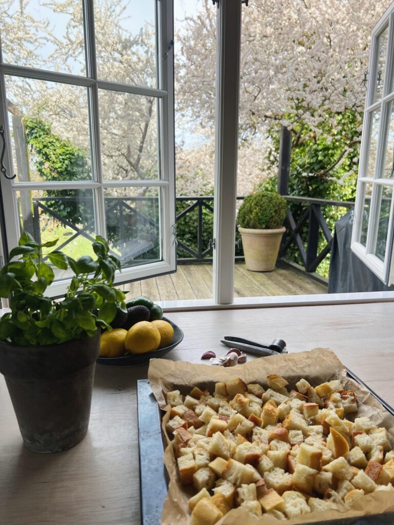 surdejsbrød fra il fornaio skåret i tern. Ligger i en bradepande klar til at komme i ovnen, så de kan ristes til sprøde brødcroutoner. I baggrunden ses et åbent vindue med udsigt til grønne planter og et blomstrende træ.