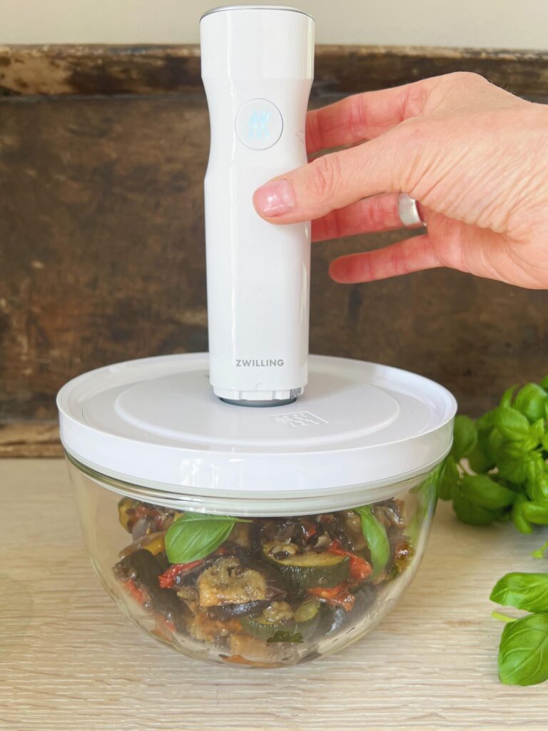 En glasskål med rester af ratatouille-måltidssalat er dækket med vakuumlåg. En hånd holder en hvid vakuumpumpe fra Zwilling over ventilen. Friske basilikumblade ligger ved siden af skålen.