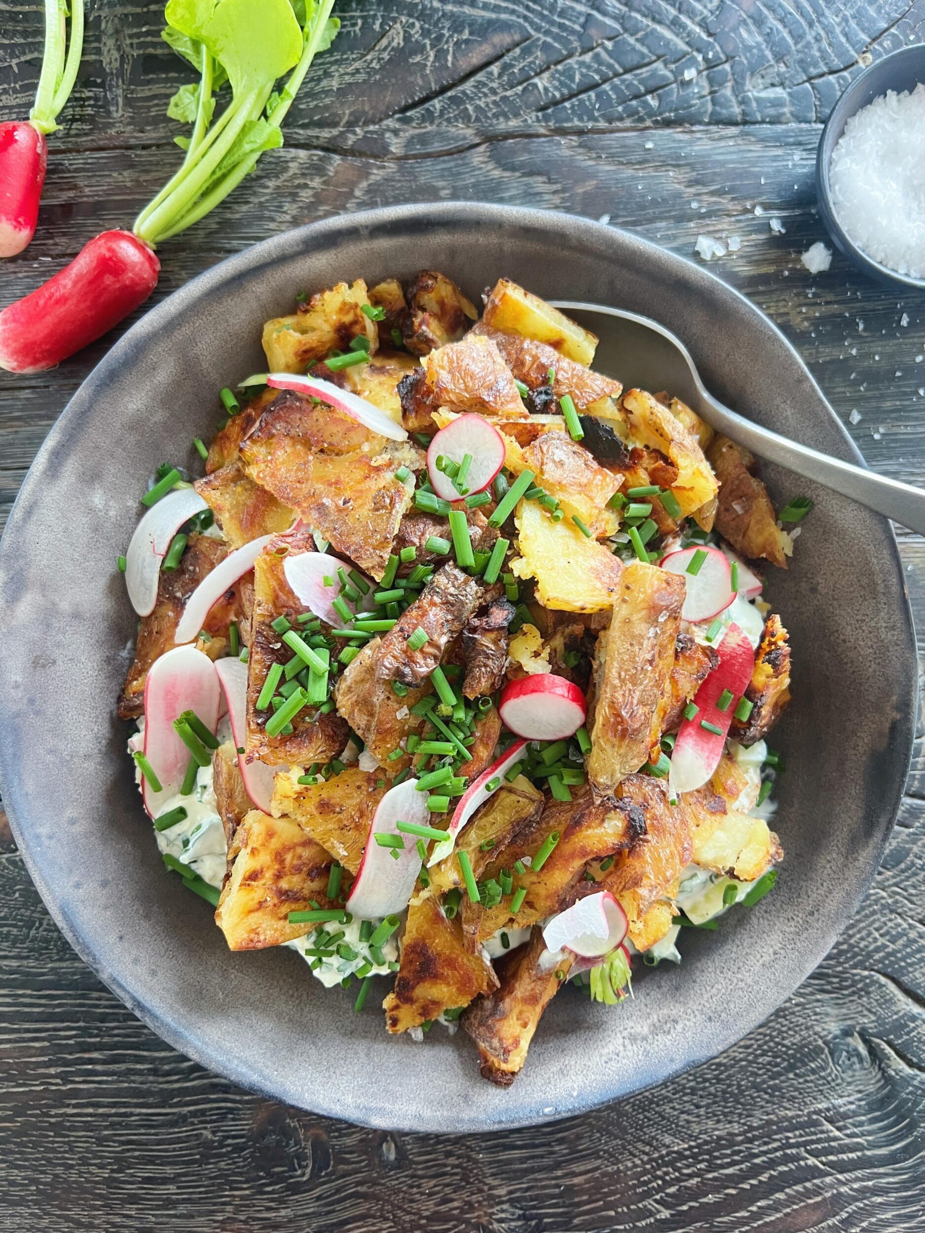 kartoffelsalat med sprøde ovnbagte kartofler. Salaten ses serveret i sort skål på sort træbord. Der ligger to radiser ved siden af. Salaten er toppet med grove stykker sprød kartoffel, snittet radisse og purløg.
