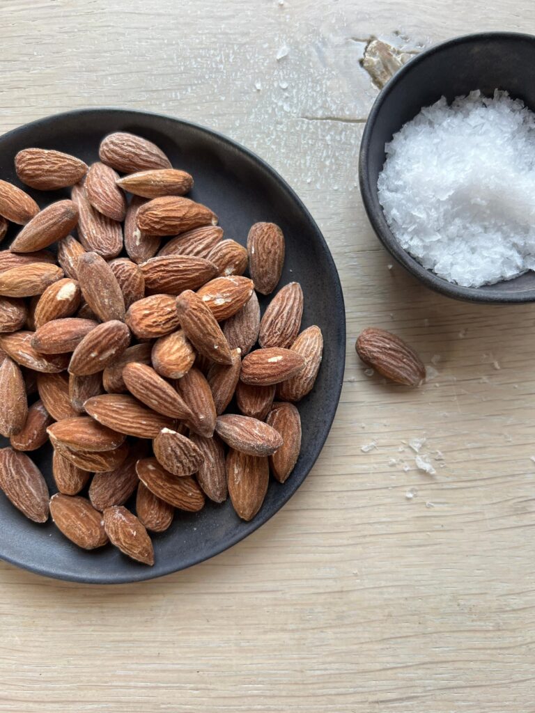 Hele saltristede mandler i en sort keramikskål. En lille sort skål med flagesalt ses ved siden af, og der ligger lidt salt spildt på det lyse bord.