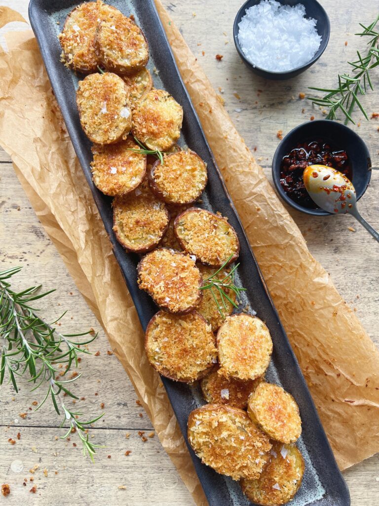Sprøde kartoffelskiver med gylden pankoskorpe på et mørkt fad ved siden af en skål crispy chili oil med en ske.